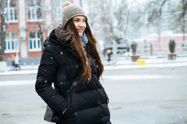 Милая длинноволосая девушка в теплой черной куртке и шляпе ходит по зимнему городу, улыбаясь — стоковое фото
