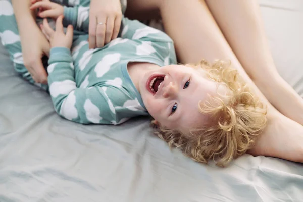 Malé dítě světlovlasý chlapec v modrém pyžamu smích, ležící na posteli vedle máma — Stock fotografie