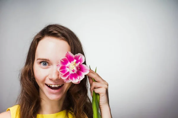 노란 드레스 포즈에 재미 있는 파란 눈 여자 얼굴에 향기로운 분홍색 꽃을 보유 — 스톡 사진