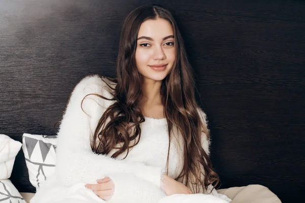 Piękna dziewczyna długowłosy w piżamie biały siedzi na łóżku, odpoczynku i chce spać — Zdjęcie stockowe