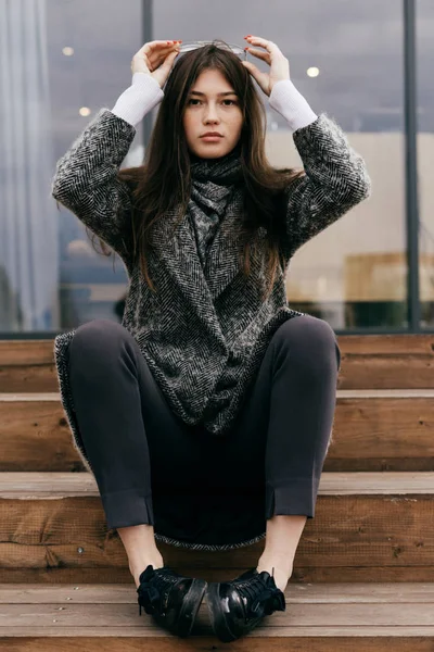 Young dark-haired girl in gray coat is resting on the steps, street style and fashion — Stock Photo, Image