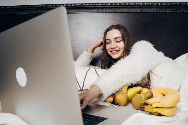 Vrolijke jong meisje ligt op het bed, ontspant en grappige ilm kijkt op haar laptop, eet fruit — Stockfoto