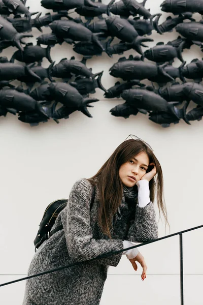 Beautiful dark-haired girl posing against the background of an unusual wall, in a gray fashion coat — Stock Photo, Image