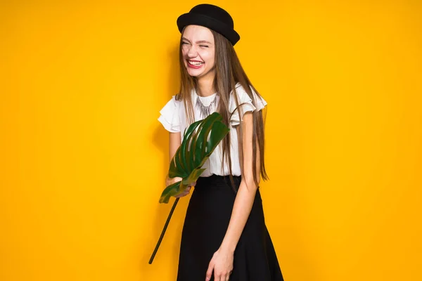 Riéndose elegante modelo de niña en sombrero negro posando sobre fondo amarillo de la pared, sosteniendo una hoja verde en las manos — Foto de Stock