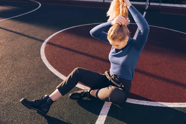 Kék pulóver-karcsú szőke lány ül a földön egy sportpálya — Stock Fotó