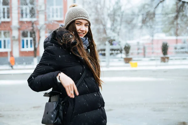 Szczęśliwy, że długo haired Dziewczyna w ciepły czarny płaszcz i kapelusz idzie przez miasto zima, jest zimno — Zdjęcie stockowe