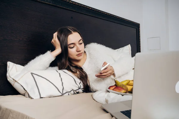 Giovane ragazza si trova a letto sotto una coperta bianca, a riposo, mangiare frutta e guardando nel suo smartphone — Foto Stock