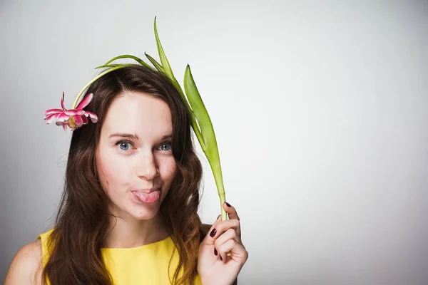 Funny blue-eyed dívka v žlutých šatech ukazuje jazyk a má květ — Stock fotografie