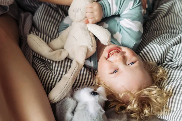 Little blond haired curly child boy in blue pajamas plays, laughs, lies on the bed — Stock Photo, Image