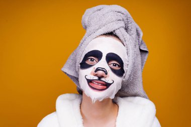 sevimli kız gülümsüyor, yüzünde bir panda yüz nemlendirici maske başında beyaz bir havlu ile