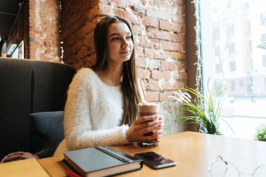 sevimli uzun saçlı kız beyaz kazaklı bir kafede oturuyor, lezzetli kahve içer ve pencereden görünüyor