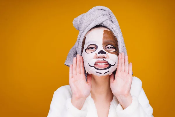 Cheerful girl with a towel on her head wants to be beautiful, on her face a moisturizing mask with a dog's face — Stock Photo, Image