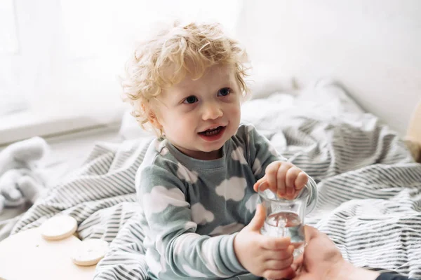 Legrační blond vlasy kudrnaté chlapeček sedí na posteli a úsměvy, nápoje voda — Stock fotografie