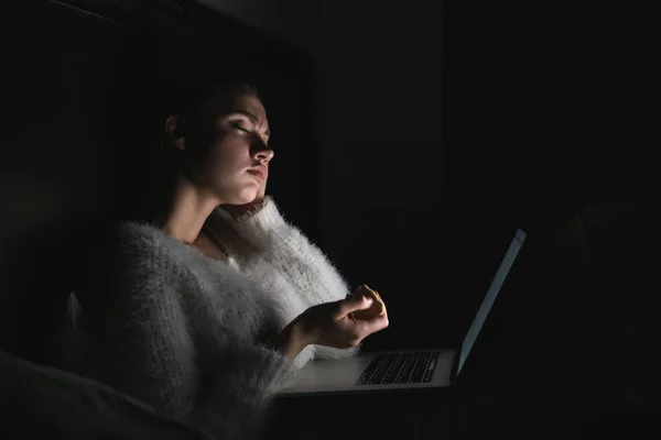 Уставшая молодая девушка, сидящая вечером в постели и смотрящая скучное кино на ноутбуке — стоковое фото