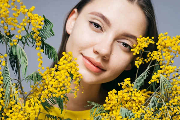 Encantadora hermosa chica con la piel limpia sostiene un ramo de mimosa amarilla, sonríe y mira a la cámara — Foto de Stock