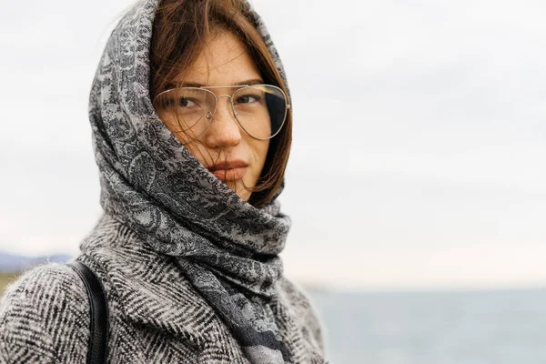 Красива дівчина брюнетка в сірому шарфі і окулярах насолоджується видом на море і похмурою погодою — стокове фото