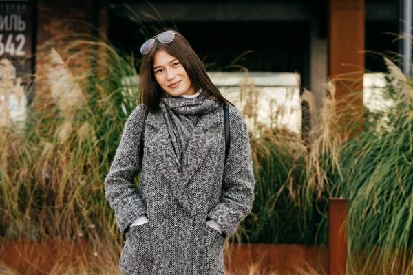 Confiant belle fille aux cheveux bruns en manteau gris à la mode posant dans les rues de la ville, souriant — Photo