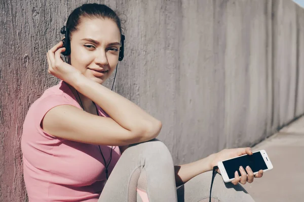 Fin smal flicka i en rosa tröja vila efter ett långt träningspass, lyssna på musik i hörlurar — Stockfoto