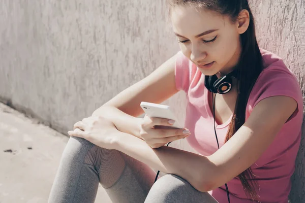 beautiful slender girl in a pink t-shirt sits on the ground, looks into her smartphone, neck hangs on her neck