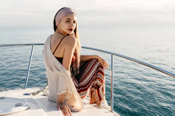 Kedves fiatal lány pózol egy jachton, szemben a tenger és a naplemente, elegáns ruhákat mosolyogva — Stock Fotó
