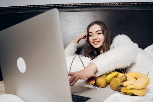 Красива мила дівчина лежить в ліжку дивитися смішний фільм на ноутбуці і їсти фрукти — стокове фото