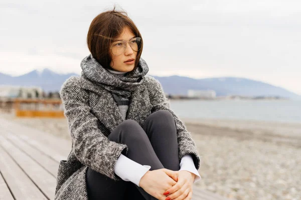 Young brunette girl in gray coat enjoying cloudy weather and sea scenery — Stock Photo, Image