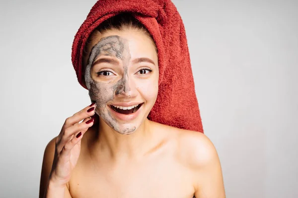 Een grappig meisje met een rode handdoek op haar hoofd een klei masker toegepast op de helft van het gezicht — Stockfoto