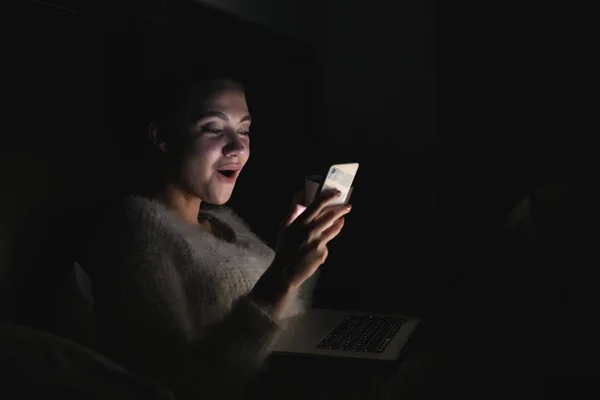 Divertente ragazza seduta a letto a tarda notte, guardando il telefono e ridendo — Foto Stock