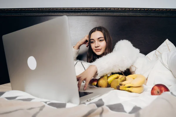 Jong meisje ligt op een bed in de avond, charmant eet vruchten en een reeks kijkt op een laptop — Stockfoto