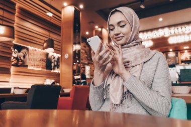 Onun smartphone üzerinde mesaj yazarken güzel Arap kız şirin bir kafede oturan bir başörtüsü ile