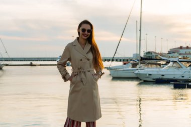 güneş gözlüğü ve gün batımında liman içinde poz bej bir palto çekici uzun saçlı kız