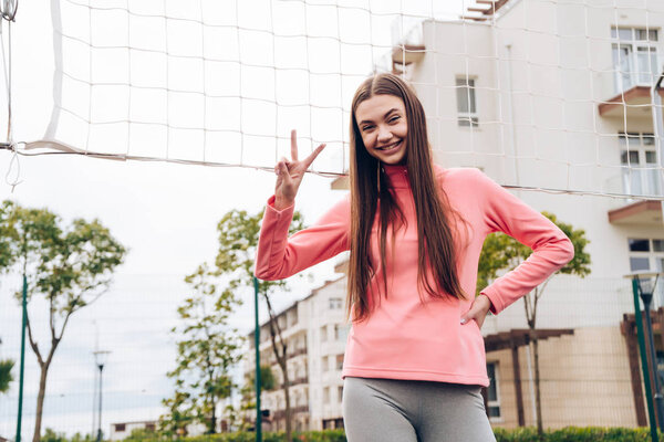 веселая активная девушка отдыхает после игры в волейбол на спортивной площадке, улыбается и показывает два пальца
