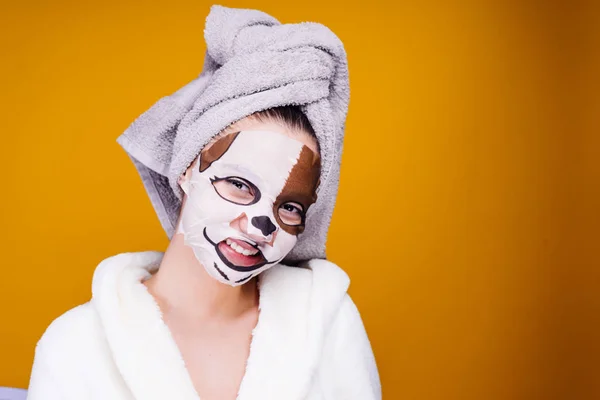 Une fille drôle dans une robe de chambre éponge blanche sourit, un masque hydratant avec le visage d'un chien sur son visage, une journée de spa — Photo