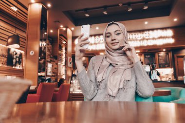 bir başörtüsü başında büyüleyici bir Müslüman kızla rahat restoranda oturur ve ona yiyecek için bekler, bir akıllı telefon tutar