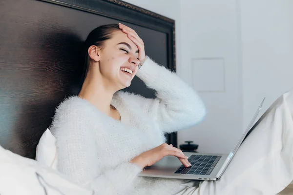 Chica joven en pijama blanco se sienta en la cama por la noche, se ve divertido video en su computadora portátil —  Fotos de Stock