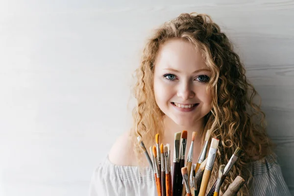 curly beautiful young woman painter holding brushes for drawing in hands, smiling