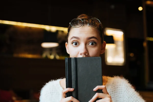 Милая удивленная девушка в белом свитере сидит в кафе, держит черную тетрадь в руках, выглядит удивленной — стоковое фото