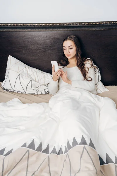 Joven chica cansada está acostada en la cama tarde en la noche, descansando, mirando su teléfono inteligente — Foto de Stock