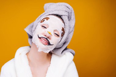 kafasını ve beyaz bornoz havlu ile neşeli bir kız spa uygulamaları, yüzünde bir nemlendirici maske sahiptir