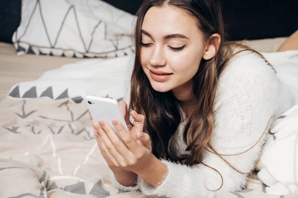 Schattig slaperig meisje in witte pyjama ligt in bed, schrijft een bericht op haar smartphone, glimlacht — Stockfoto