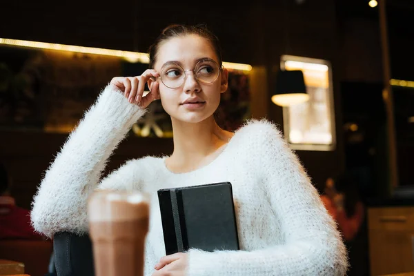 Задумчивая молодая девушка-фрилансер в очках сидит в кафе, держит в руках блокнот и думает о работе — стоковое фото