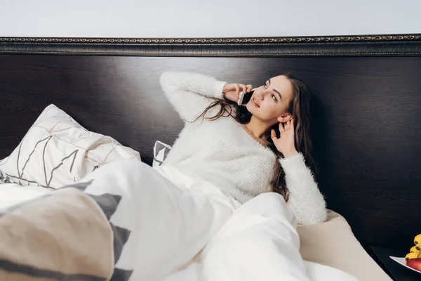 Sleepy meisje liggend in bed onder een witte deken, praten over de telefoon — Stockfoto