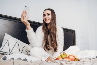 Onun smartphone bakarak uyumak hazırlanıyor genç kız beyaz pijama yatak, oturma gülümseyen