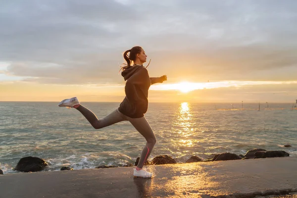 Activa chica rápida en ropa deportiva corre a lo largo del terraplén del mar al atardecer, escucha música en los auriculares — Foto de Stock