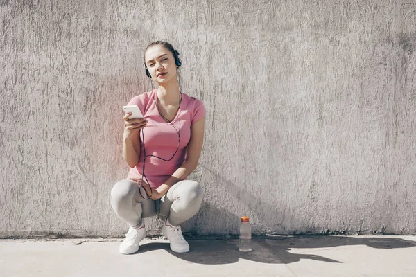 Slank jong meisje in een roze t-shirt rust na het joggen buiten, luisteren naar muziek op koptelefoon — Stockfoto