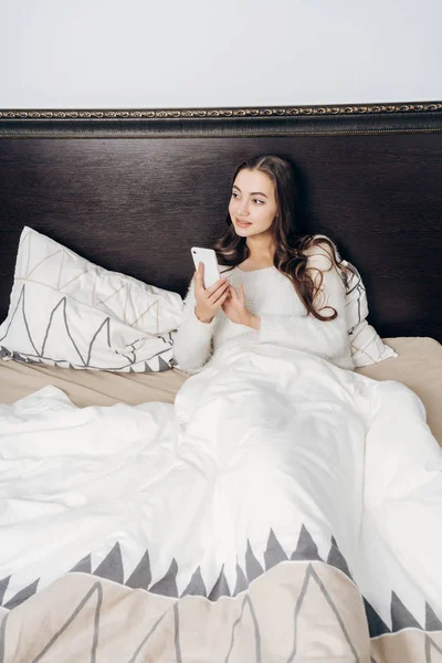 Una joven de pelo largo yace en la cama debajo de una manta blanca, sostiene un teléfono, quiere dormir — Foto de Stock