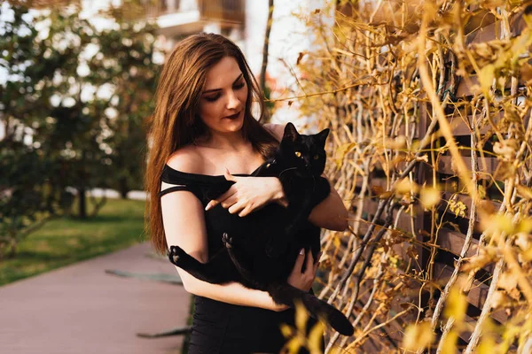 Charmant langharige mooie vrouw in zwarte jurk heeft een zwarte kat op haar handen, wandelingen in de tuin in de avond — Stockfoto