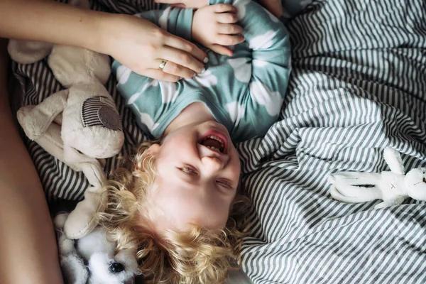 Un pequeño chico de pelo rubio en pijama azul yace en la cama junto a su madre, se ríe, se divierte — Foto de Stock