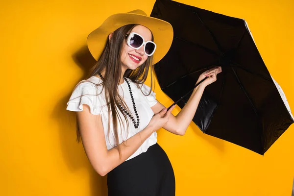 Sorridente confiante modelo menina de cabelos longos em chapéu e óculos de sol mantém guarda-chuva e posando no fundo amarelo — Fotografia de Stock