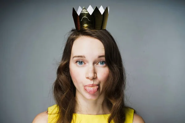 Engraçada menina de olhos azuis em um vestido amarelo mostra uma língua, uma coroa de ouro em sua cabeça — Fotografia de Stock